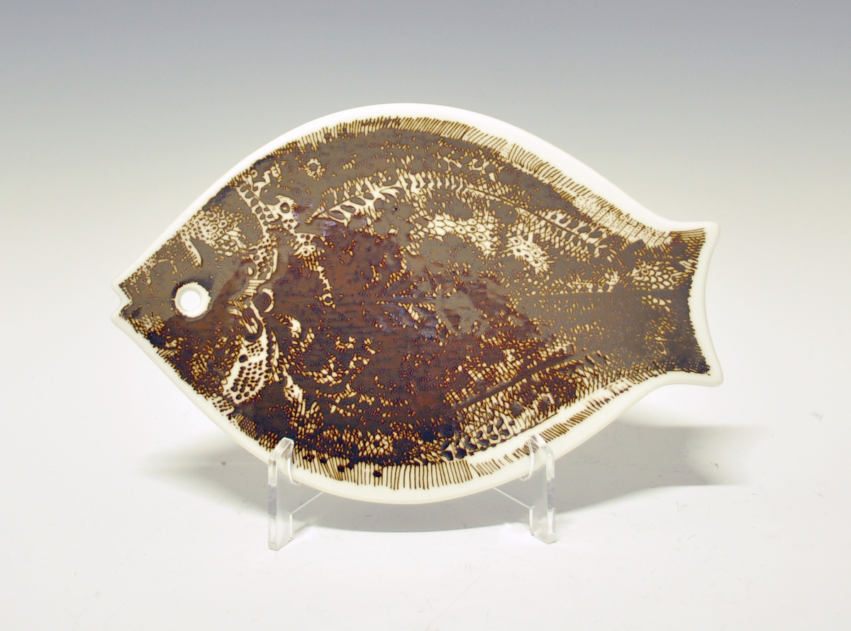 Smørefjel i porselen. Formet som fisk med hull til oppheng. Dekorert i brunt.
Design: Arne Lindaas.