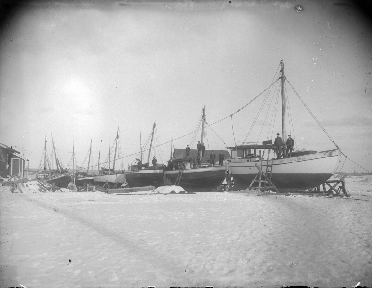 Seks seilskøyter på slipp i et vinterlandskap, trolig på Andenes. På de forreste tre båtene står menn oppstilt på dekk.