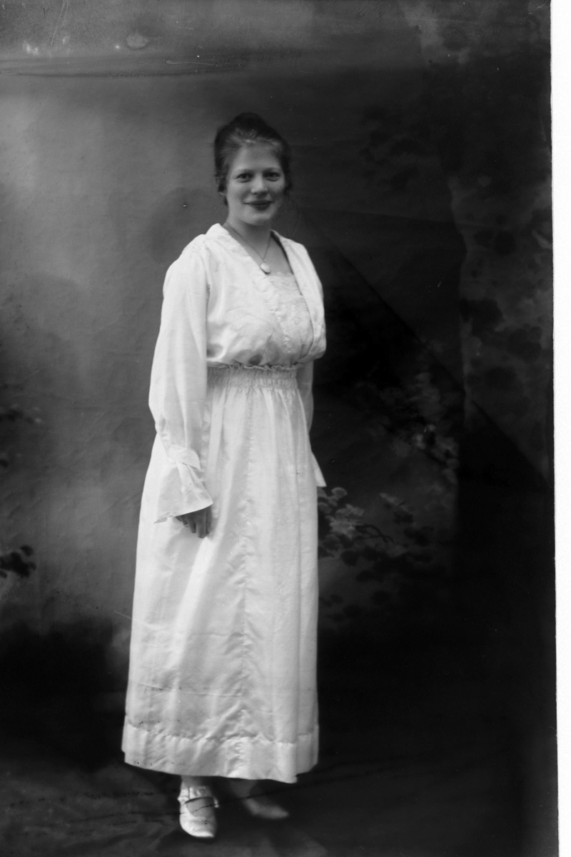 Studioportrett i helfigur av en kvinne i hvit kjole.