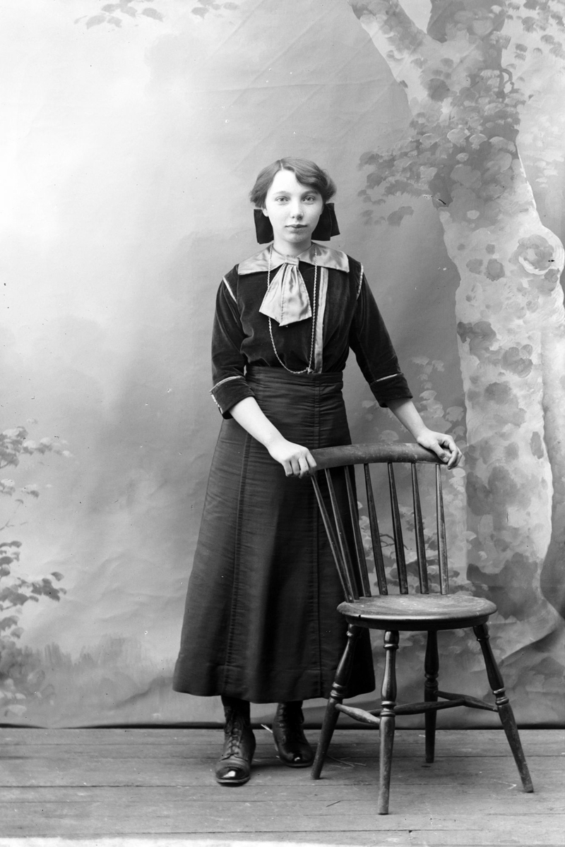 Studioportrett av en kvinne med henderne på en stolrygg.