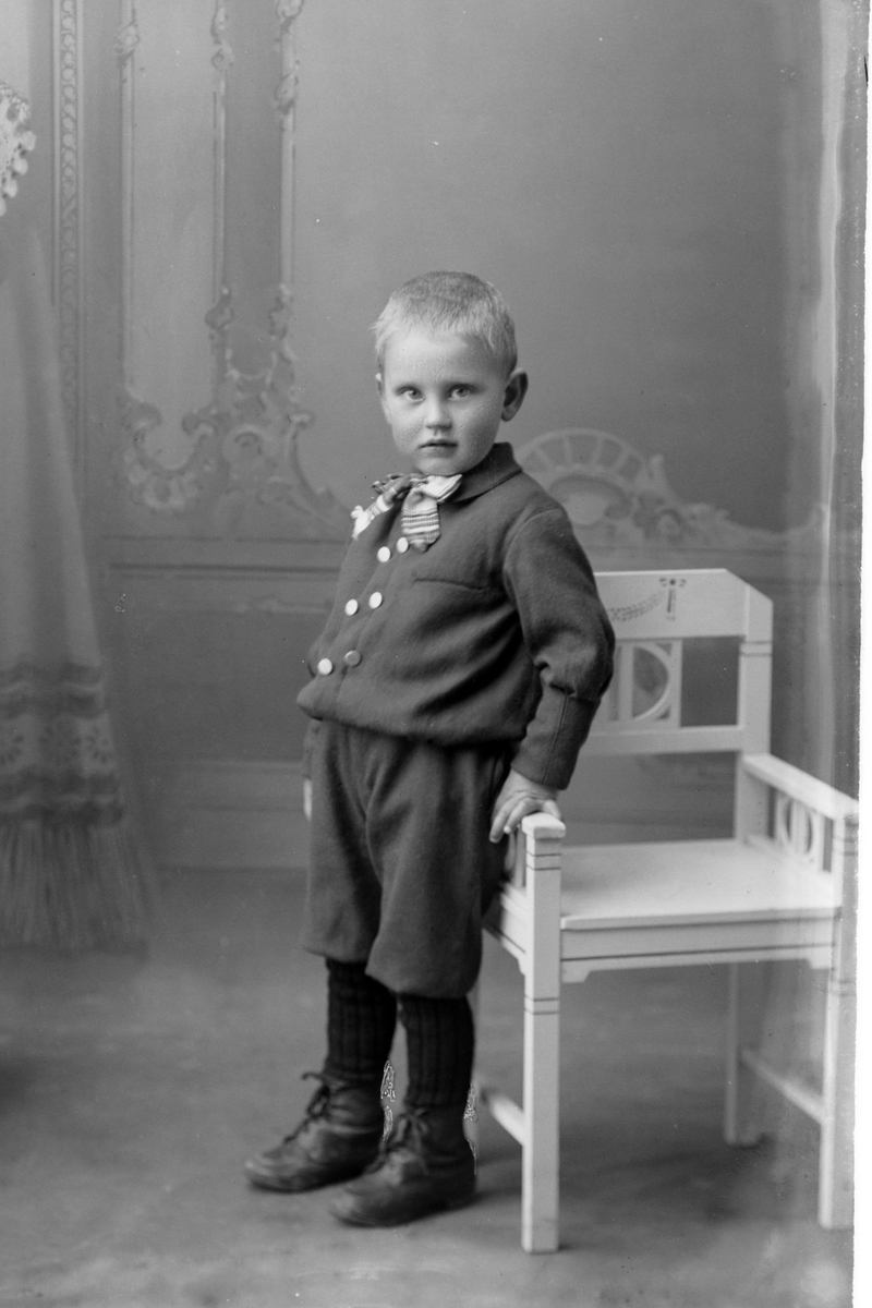 Studioportrett av en liten gutt som står ved siden av en stol.