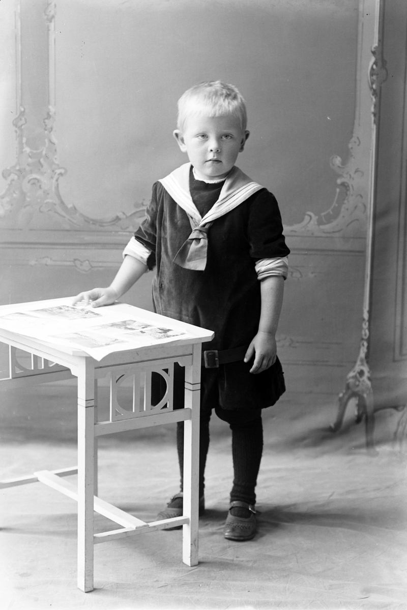 Studioportrett av en liten gutt stående ved et bord.
