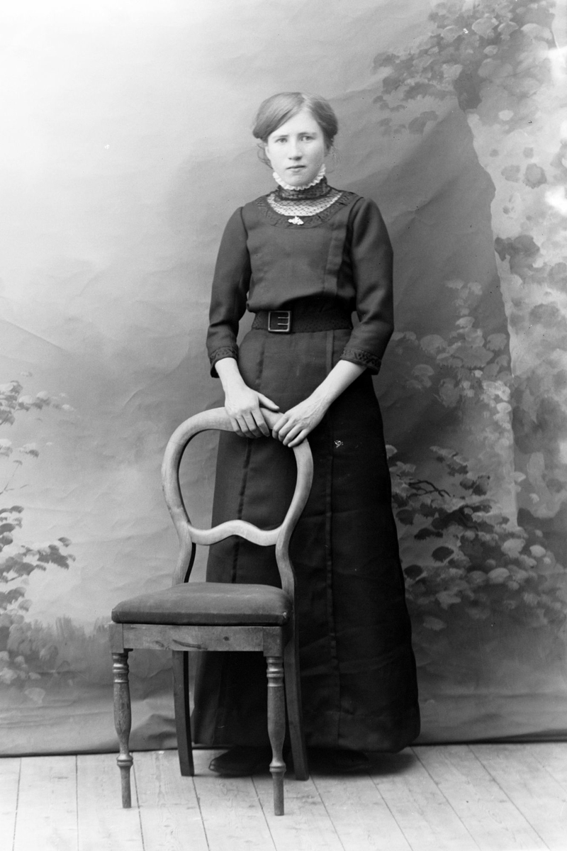 Studioportrett av en kvinne stående bak en stol.