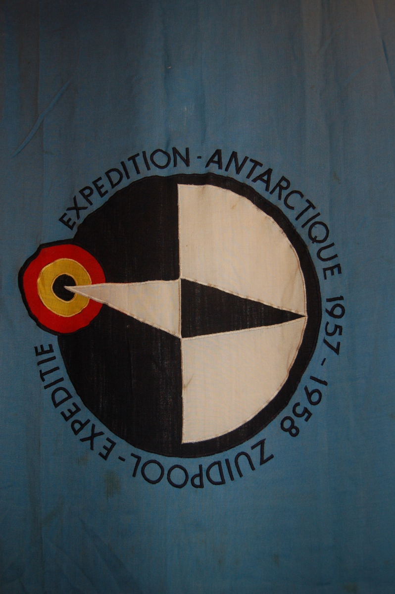 Rektangulært flagg med svart/kvit kompassnål peikende mot syd på blå botn, der syd er avmerka som en blink i rødt, gult og ssvart.