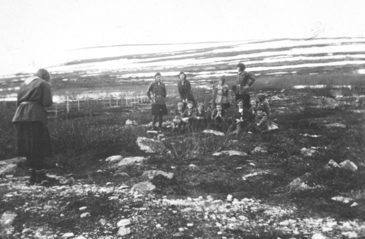 Speidertur overfor Vadsø 1933. En gruppe speiderjenter posere for ei annen speiderjente som tar bilde av dem. I bakgrunnen ses fjellet hvor det ligger snø. Naturen rundt dem består av stein og kratt
