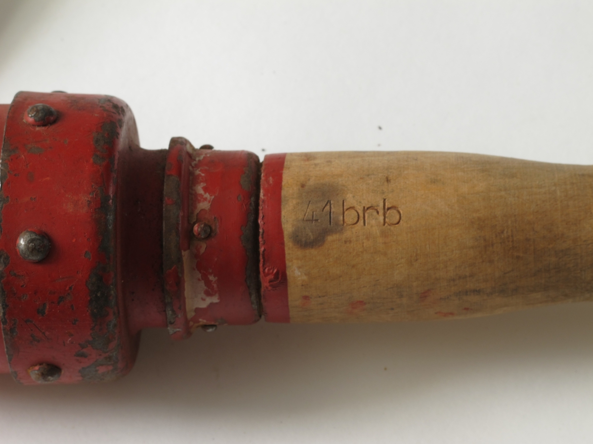 En kort rødmalt sylinder av metall, festet til et dreid skaft av tre. Metallbeslag også i skaftets ende. 