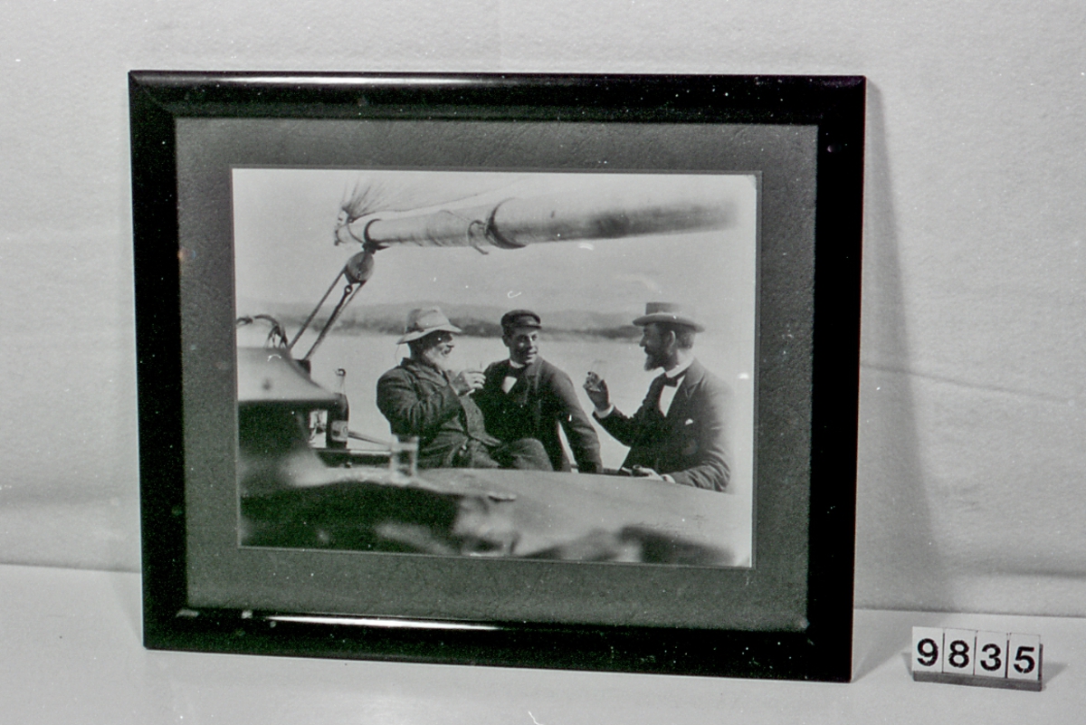 Tre menn ombord i en seilbåt. Sitter med glass i hendene.Flaske på et bord t.v