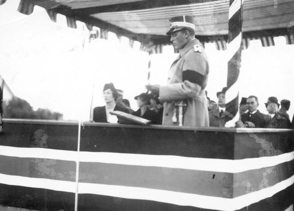 Kong Olav taler fra en tribune ved Haraldsstøtta. I bakgrunnen sees kronprinsesse Märtha og endel andre notable personer. Tribunen er pyntet med norske farger.