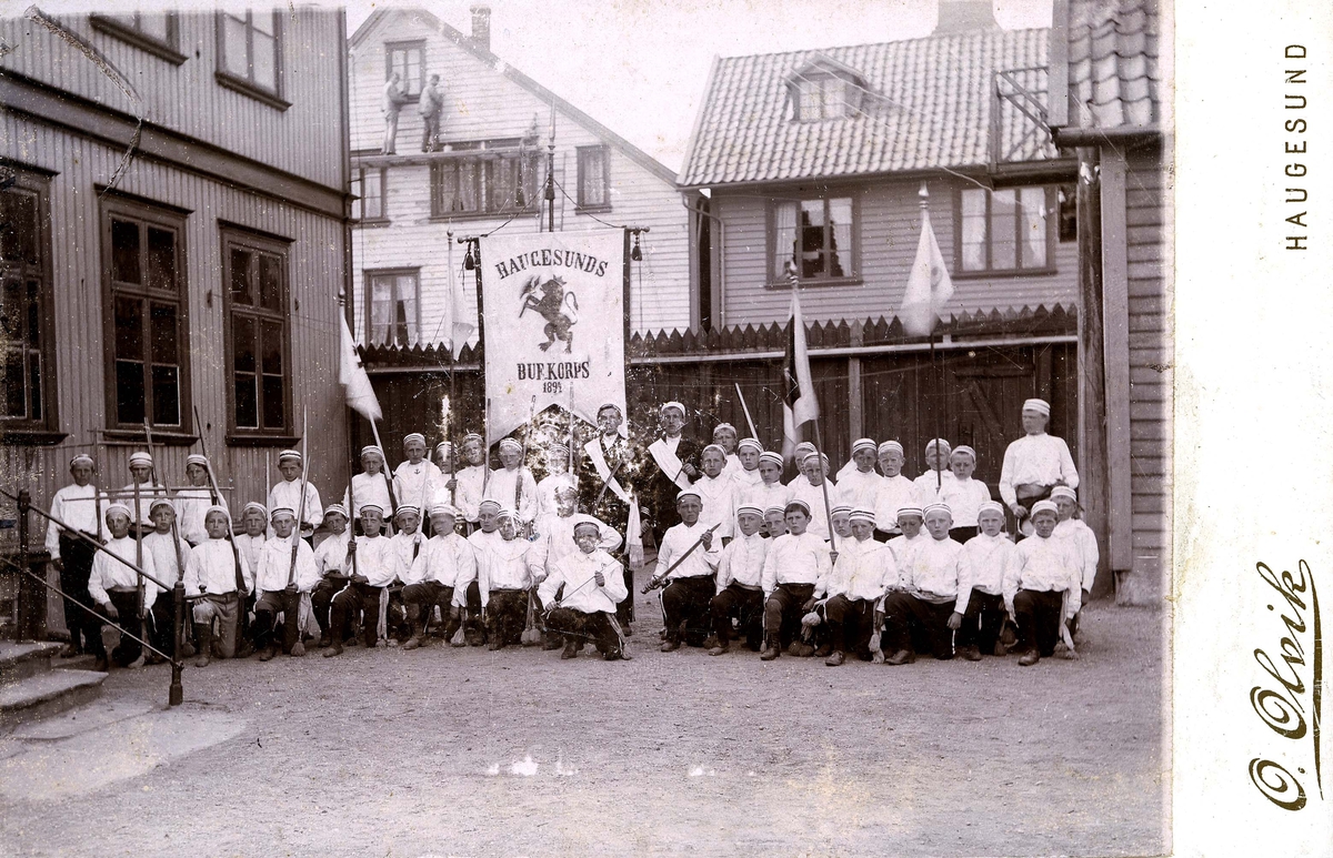 Haugesunds Buekorps med uniformer og fane. Bygårder og to menn på stillas i bakgrunnen.