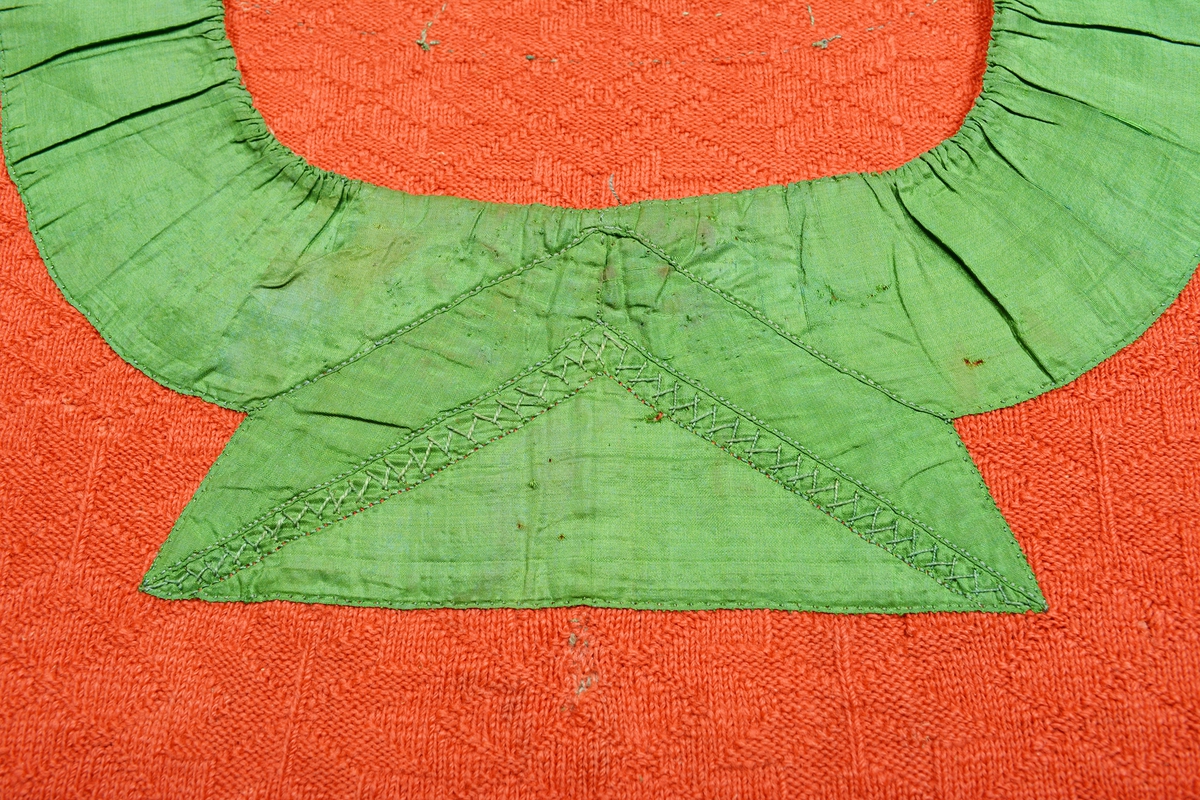 Spedetröja i rött ullgarn garnerad med ett brett grönt band i halsringningen och på bröstet. Tröjan är mönsterstickad med ett stjärnmönster över hela ytan. En avigstickad rand löper nedifrån och upp längs sidorna på både fram- och bakstycke. Litet sprund i sidorna. Det gröna sidenbandet runt halsringningen är 60 mm brett, på bröstet är bandet broderat med en V-linje i kråkspark/gles flätsöm i grönt. Halsringningen bak är fodrad med en 20 mm bred remsa i halvblekt linne. Nederdelen på ärmen är kantad med ett smalt band i rosa och grönt mönster på svart botten. Av färgskillnad/mindre blekning kan man se att nederdelen på ärmen  tidigare haft ett bredare påsytt band. Sprundet på ärmen är 50 mm långt, knäppning med knapp och knapphål. Knappar saknas, langetterade knapphål i grönt. Banden är påsydda för hand.Masktäthet/cm: 5 maskor på höjden och 4 maskor på bredden. Axelbredd: 220 mm. Ärmlängd: 455 mm. Bredd nedtill: 410