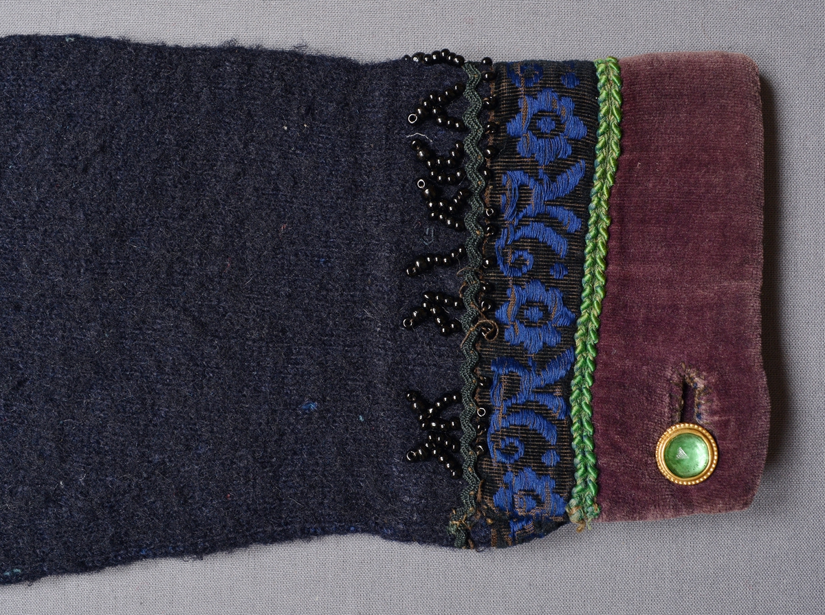 Spedetröja i mörkblått ullgarn garnerad med band, tyg och pärlor i halsringning och vid nederdelen på ärmen. Tröjan är slätstickad med mönsterstickning på axeln och en rätstickad rand i nederkanten. Litet sprund i sidorna. Lila sammetskant (35 mm bred) runt hals och och nederdelen på ärmen, bredvid denna ett smalt grönt garneringsband, sedan ett mönstrat sammetsband i blått på svart botten och ytterst ett svart band med påsydda svarta pärlor.Ärmsprundet hålls ihop av en gulmetallknapp med en infattad grön, facetterad glaspärla, handsydda knapphål.Mitt bak är halsringningen delad av ett 90 mm långt sprund som knäpps med hyska och hake, sprundet är också kantat med lila sammet. Invändigt är halsringning och ärmkant fodrade med halvblekt linne. Foder, band och pärlor är påsydda för hand. Tröjan är valkad.Masktäthet/cm: 4 maskor på höjden och 3,5 maskor på bredden. Axelbredd: 170 mm. Ärmlängd: 420 mm. Bredd nedtill: 340 mm. Märkt invändigt bak vid halssprundet med påsydd tyglapp med texten: "N° 165. a".
