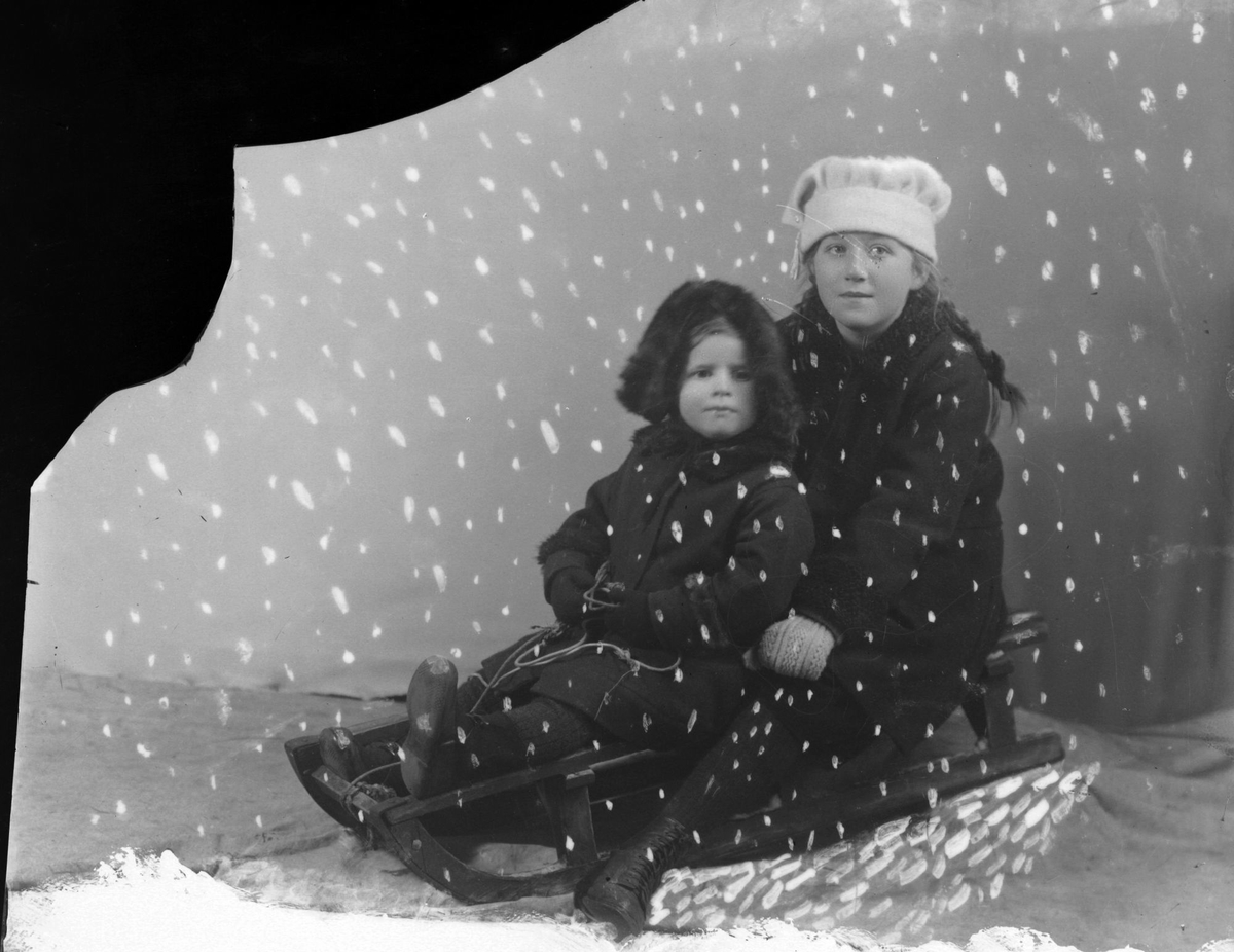 Studioportrett av to barn på en kjelke, med kunstig snø lagt på bildet.