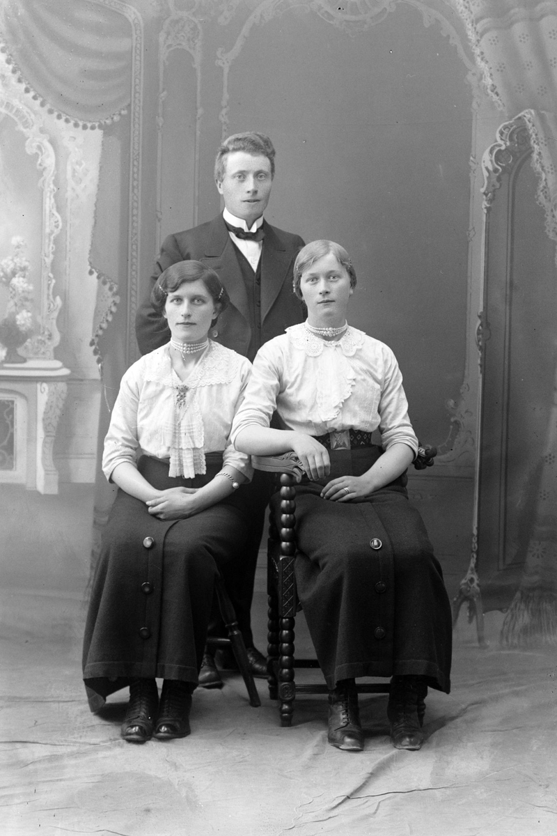 Studioportrett av en mann stående bak to kvinner som sitter.