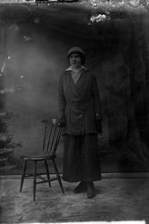 Studioportrett av en kvinne stående ved en stol.