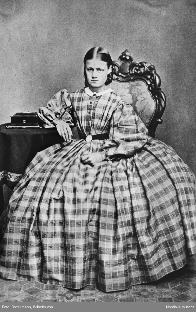 Porträtt av ung kvinna, Anna Carolina Östrand (född 1845) sittande vid bord iklädd rutig klänning, krinolin, med puffärmar och vit krage.