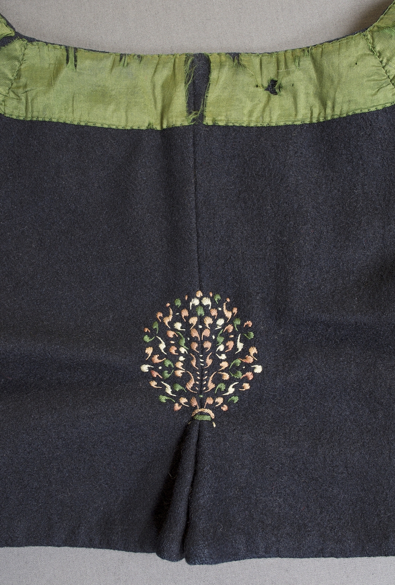 Blå vadmalströja garnerad med band och broderier. Tröjan har en rund öppning fram på bröstet och skört bak. Runt halsringning och öppningen fram, längs nederkanten på framstycken och ärmar är tröjan dekorerad med ett grönt 38 mm brett sidenband. Nederkanten på ärmen är dessutom dekorerad med ett mönstrat band i rosa och grönt silke på svart sammetsbotten. Framstyckena är också prydda med broderier på och intill det gröna bandet. Broderat blommönster finns också ovanför bandet på ärmen samt ovanför skörtet mitt bak. Framtill knäpps tröjan på två ställen med en hyska och hake. Ärmlängd: 480 mm. Ärmen är sydd i ett stycke samt en kil. Ärmsprundet är 80 mm långt och knäpps med en rund bärnstensknapp, tränsat knapphål. Axelbredd: 120 mm. Bakstycket har söm mitt bak. Tröjan är helfodrad med handvävt, tuskaftat halvblekt linne. Broderierna är utförda i plattsöm och flätsöm i olika nyanser rosa och gult samt i blått, vitt och grönt silke. Foder och band är påsydda för hand.