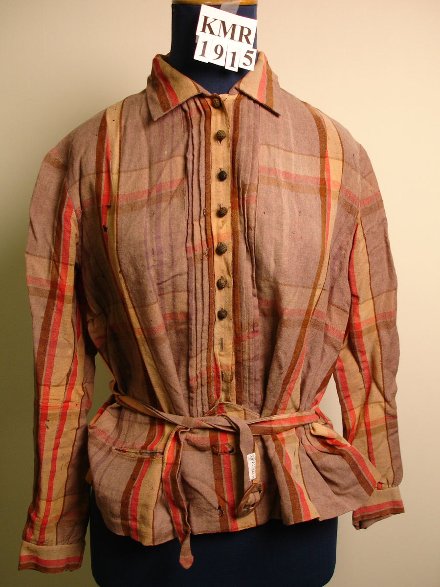Skjorte med belte til dame. Skjorten er i mange farger, med ruter og striper. Lukkes foran med brune stoffknapper. Nederst på høyre side er det en liten lomme. Stoffhemper i sidene til beltet.