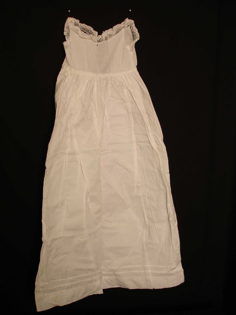 Dukkeklær, kjole i hvit bomullstoff uten erm,  blonderkant rundt hals, knepping i ryggen. Fra slutten av 1800 til begynnelsen av 1900