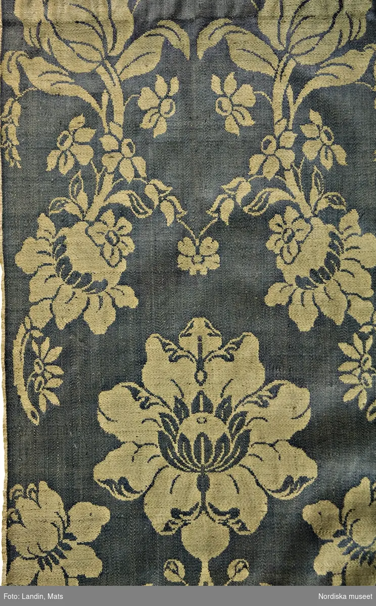 Förkläde av stormönstrad ylledamast i 1700-talsmönstring. Gulgrönt stort blommönster på mörkgrön botten. Förklädet är hopsytt av 2 hela vådbredder med stadkanter i var sida. Upptill rynkat mot en remsa av mörkblå vadmal med fastsydda knytband av olika hemvävda band. I ena sidan ett ripsband i brunt, blått och gult ullgarn på linvarp och i den andra sidan ett mönstervävt band med rött upplockat mönster på oblekt linnebotten. I mönstret är invävt initialerna L K O D. I nederkanten är förklädet skott med 3,5 cm brett ylleband skarvat av ett helrött band och ett rött med smala vita ränder. På insidan av midjelinningen broderat med vitt: 90.
Förklädet är unikt och mycket välbevarat sånär som på några malhål på midjeremsan av vadmal.
Ylledamast av denna höga kvalitet vävdes i första hand i Norwich, England, men tillverkning fanns även på andra platser eftersom detta var ett modernt tyg under 1700-talet. Till Sverige kom utländska tyger av denna sort in i allmogedräkten i slutet av 1700-talet och eftersom det var dyrbara köpetyger användes de till högtidsdräkt i första hand bröllopsdräkterna för både kvinnor och män, vanligast som livstycken och västar, i enstaka fall som kjolar och ännu mer sällan som förkläden.  I detta fall kan man se hur rapporten har sett ut, vilket är svårare på de tillskurna livstyckena. Eftersom sådana utländska tyger hade importförbud under 1700-talet kan de ha kommit illegalt via gränshandeln med Norge.
Förklädet kom in tillsammans med ett par sammanbundna kjolväskor av  rött tyg med påsydda silverband, inv.nr  98867 som använts som brudklädsel. Det kan mycket väl vara från samma brud.
/Berit Eldvik 2008-06-09

I sin bok "Möte med mode" (2014), sid 120-121, skriver Berit Eldvik att förklädet är vävt i Norwich.