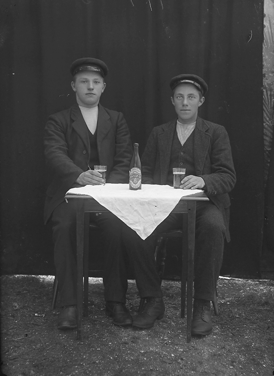 Portrett. To menn sitter ved et bord. På bordet er det en hvit duk, ei flaske med øl, 2 glass med øl. Bildet er tatt på Ramberg i Lofoten.
Fra v.: Sverdrup Lie Pettersen, f. 1895,Valle øvre og Lyder Parelius Larsen, f. 1899, Kilan.