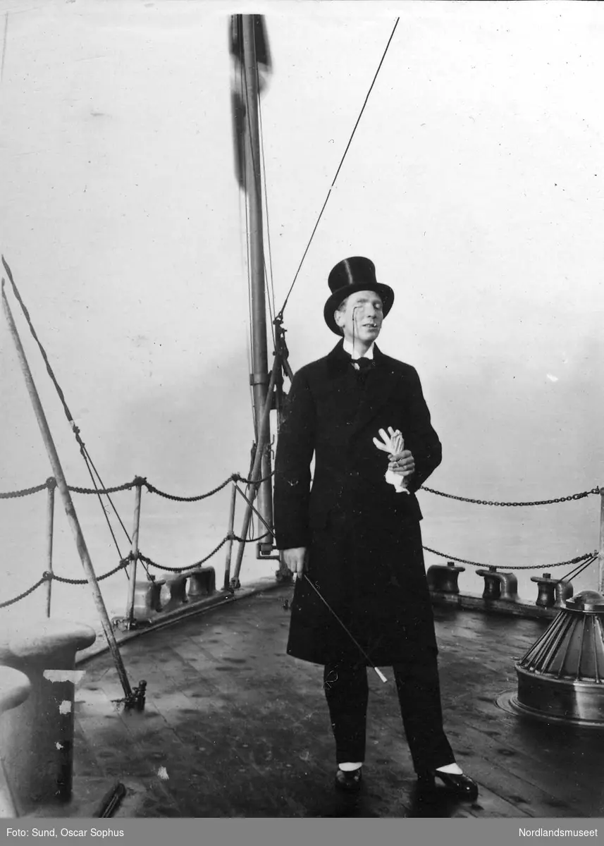Mann med flosshatt, monokkel, svart frakk, hvite hansker, stokk og høyhelte sko står forut på et skip, vi ser skipsdekk, reling, flaggstang, pullerter og ventil.