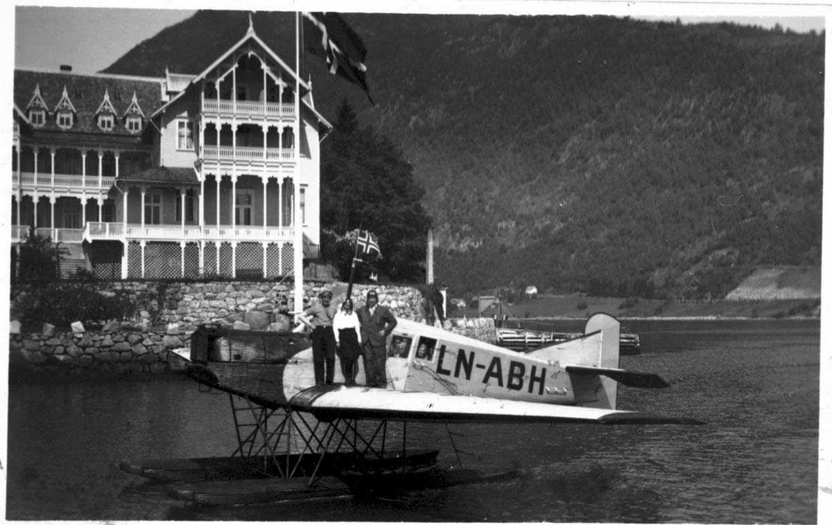 Ett fly på havet, Junkers F 13W. Senere omregistrert LN-ABH og solgt til Nord-Norges Aero A/S, Narvik. På vingen av flyet står det to menn og en kvinne. Det sitter også noen personer inne i flyet. Ei større bygning i bakgrunnen.