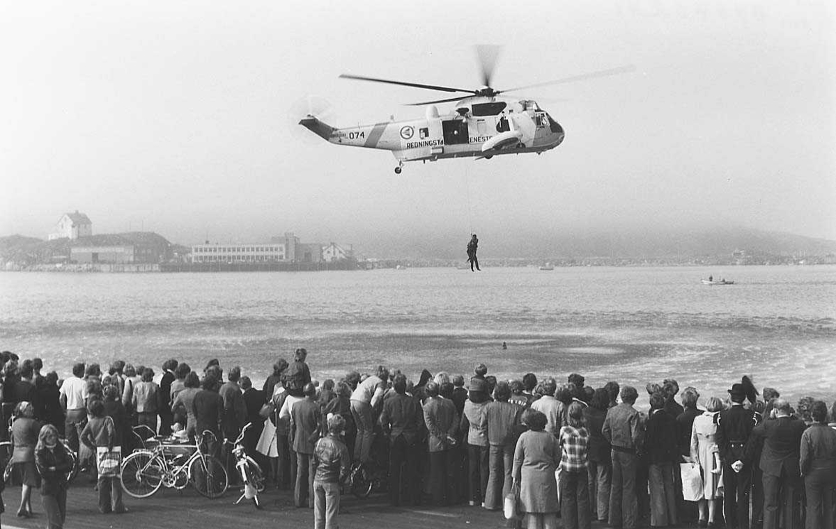 Helikopter - Sea King 074 - foretar en redningsaksjon fra sjøen. Tilskuere i forgrunnen, bygninger i bakgrunnen.