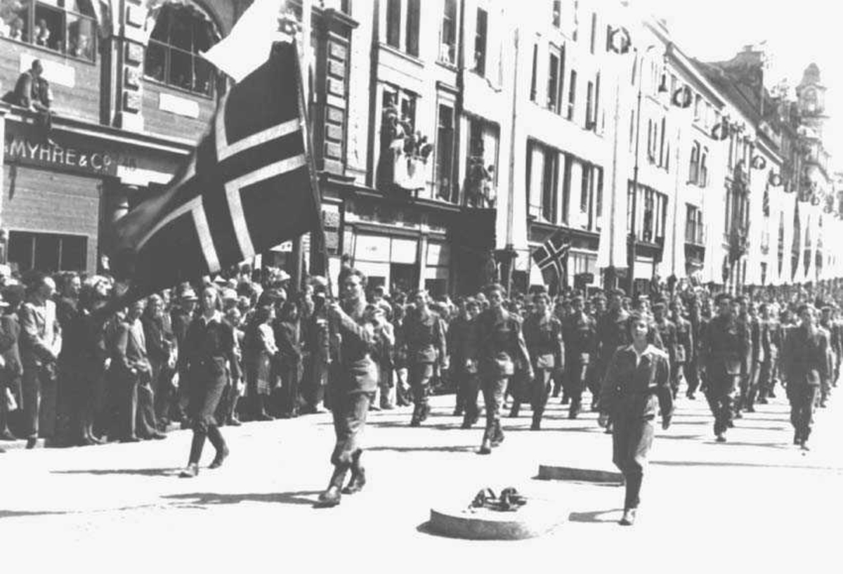 Militærtropp som masjerer i gaten. Den første bærer et norsk flagg. Folkemengde ved siden av. Bygninger i bakgrunnen.