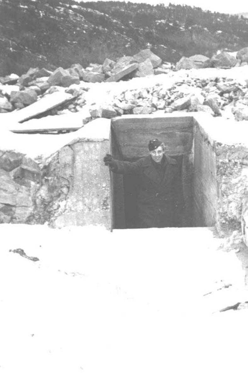 En person som står i åpningen av en bunker. Mann i militæruniform. Fjell i bakgrunnen.
