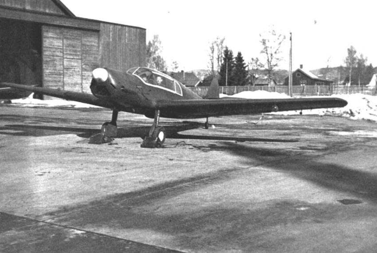 Et fly på bakken. Messerschmitt 108. Står foran en hangar. Personer inne i flyet. Bygninger i bakgrunnen.