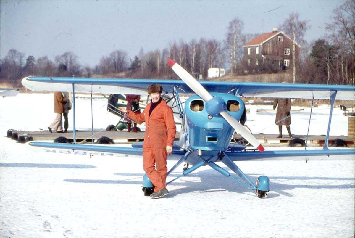 Ett fly ute på et isdekt vann, EAA Acro Sport, LN-BGK. En person, mann, står foran flyet. Flere personer på ei trekai og en bygning i bakgrunnen.