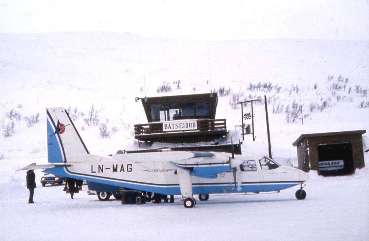Lufthavn. Ett fly på bakken, Britten-Norman BN-2A Islander, LN-MAG fra Norving. Flyplassbygninger og personer i bakgrunnen. Snø på bakken.