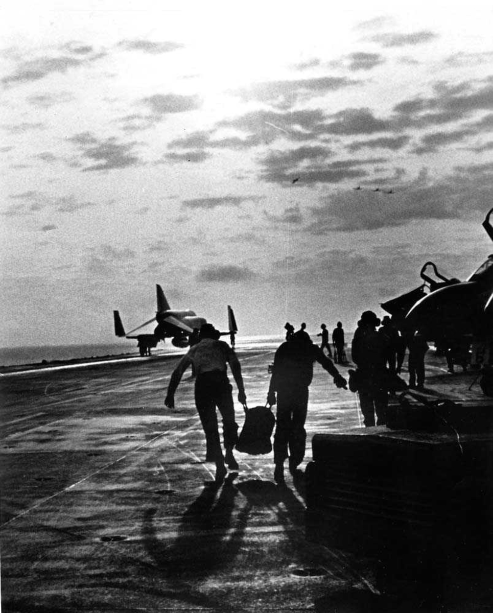 To personer på dekket til et hangarskip som bærer noe mellom seg. Flere personer og fly i bakgrunnen, F-4 Phantom.