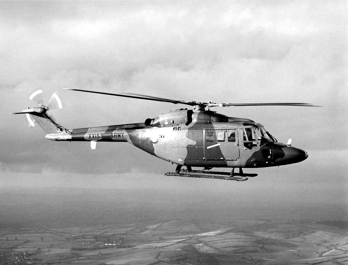 Ett helikopter i luften, over landbruksområder. Westland Lynx AH Mk.1. Halebjelke merket XX153 ARMY. Kamuflasjefarger.