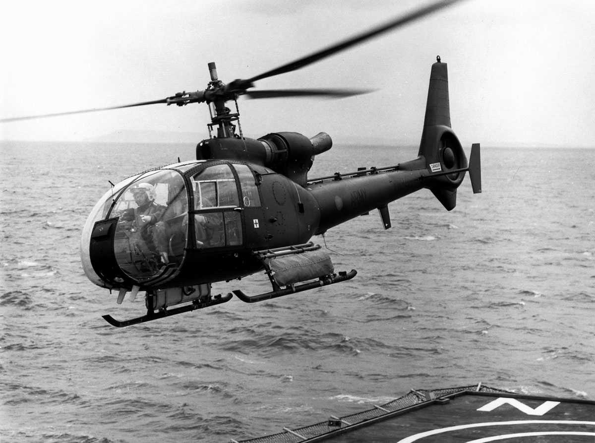 Ett helikopter i luften, like over helipad/hoveddekk på skip (nede t.h.). Aeropatiale/Westland Gazelle SA341B/AH Mk.1 tilhørende British Army merket XW848. Pilot og annenpilot ses i cockpit.