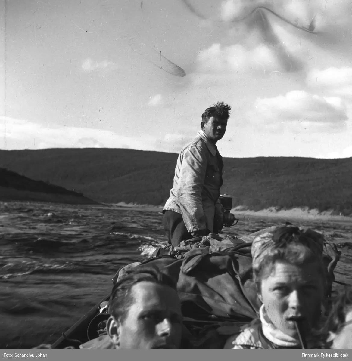 Bildet er tatt ombord på elvebåten: vi ser ansiktet til en mann og en kvinne som sitter midt på båten. Bak på båten står en mann og styrer. Bakerst fjellene.