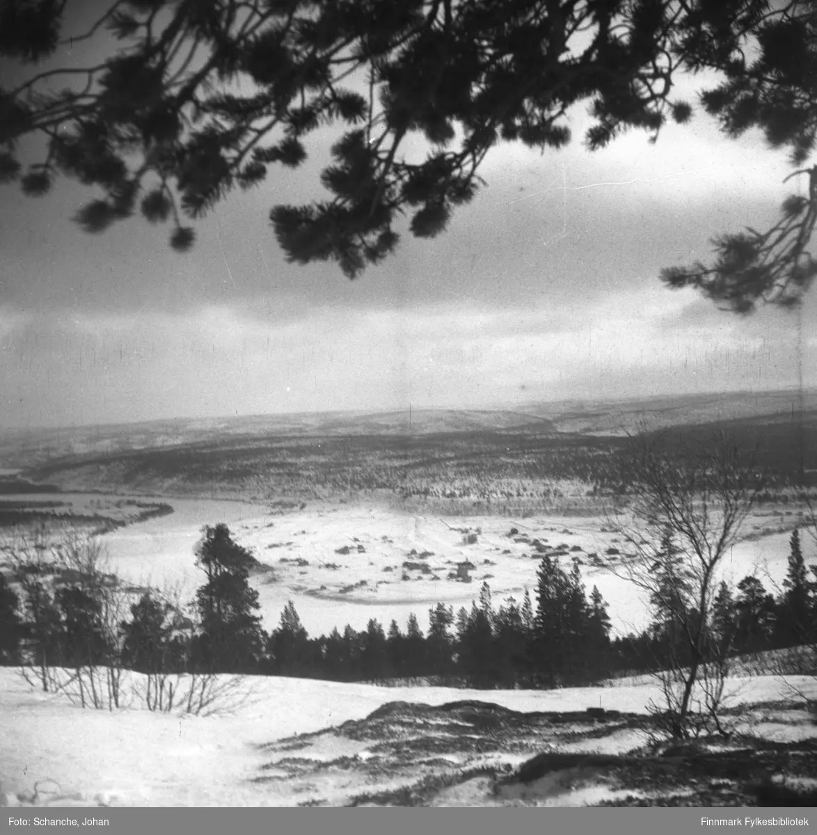 Oversiktsbilder over Karasjok tatt fra fjellet ned mot bygda i dalen.  Fotografen har stått under en stor furu, furugrein henger over landskapet på bildet.  Samme motiv som i FBib.95066-105 og 106.
