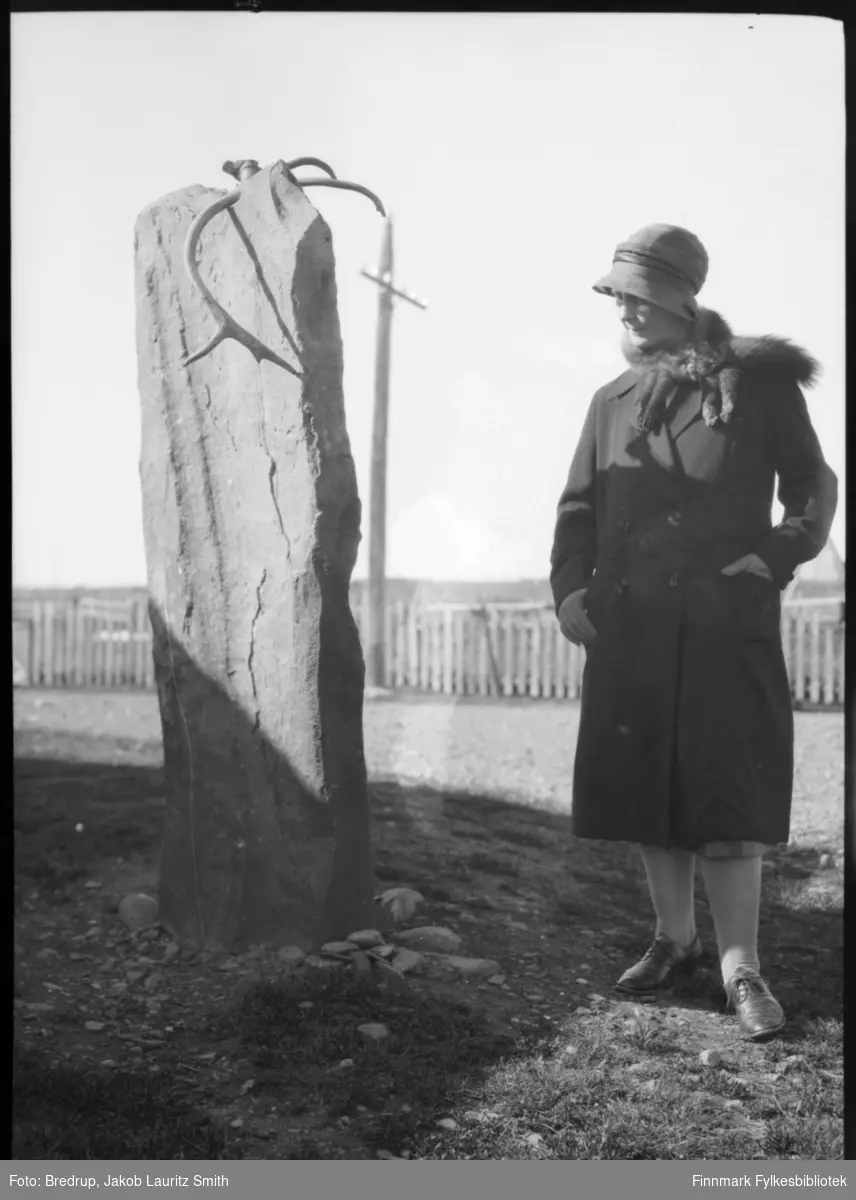 Hildur Norvik kledd i hatt og kåpe med revepels, betrakter Ivarsteinen på Sletten i Vadsø. Steinen har antakelig opprinnelig vært en samisk offerstein.