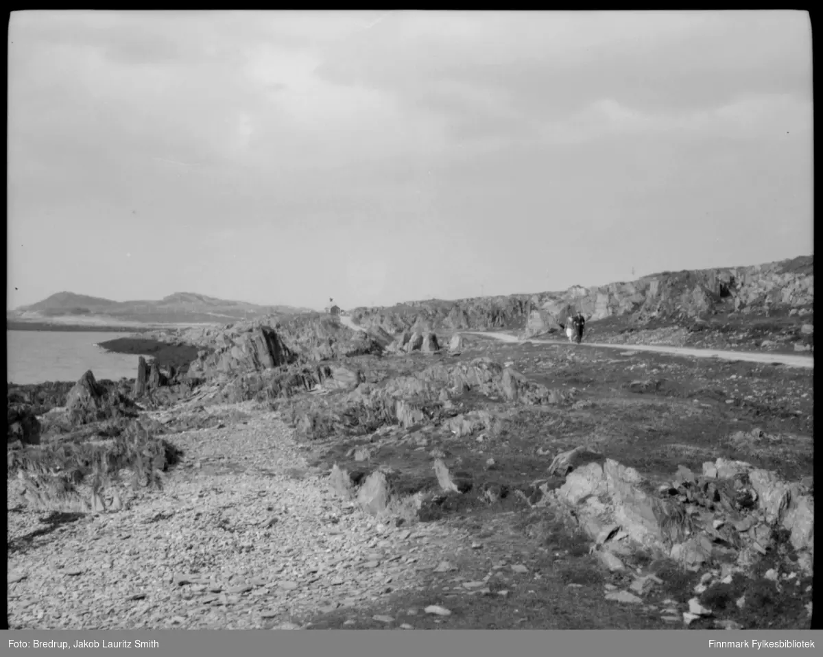Et sommerkledt par spaserer langs veien, i bakgrunnen en liten hytte.  Til venstre sjøen.  Landskapet domineres av klipper og stein. I følge vår informant, er bildet tatt i Skagen, Vardø.