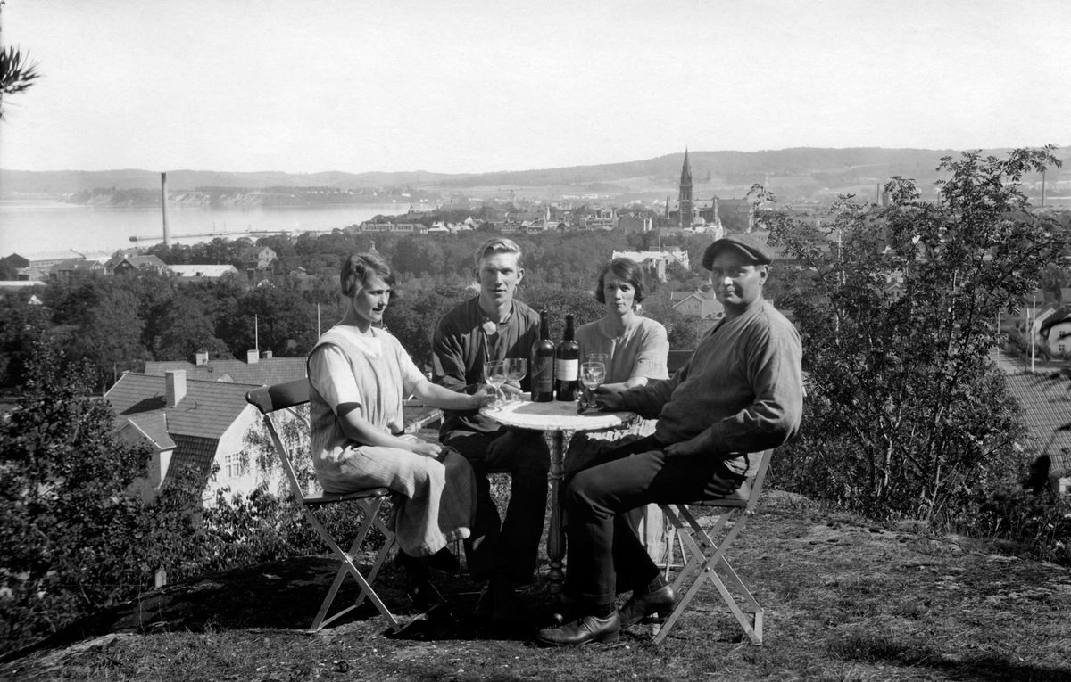 Två kvinnor och två män sitter vid ett bord med vinflaskor och glas utanför Alphyddan i Jönköpings Stadspark. I bakgrunden syns Jönköping stad och en del av Vättern.