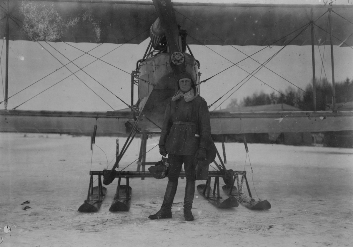 Porträtt. En militär flygare står framför flygplan Albatros BIIa med skidor på ett flygfält, vintertid. I bakgrunden syns tälthangarer.