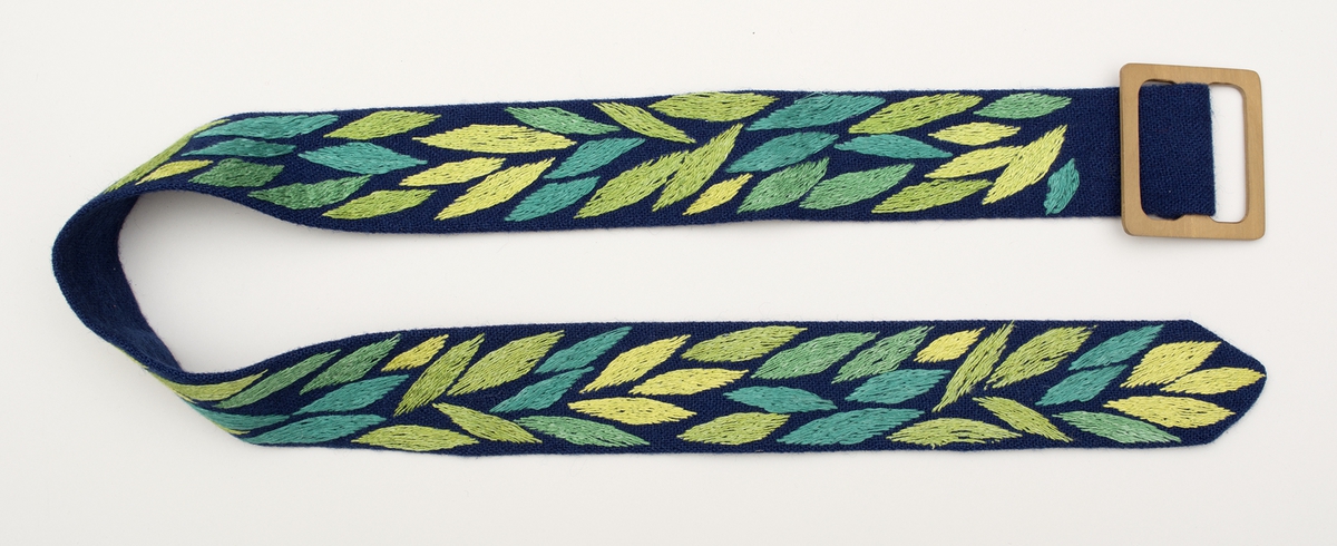 Skärp i ylletyg, broderat med olika gröna, turkosa, gröngula nyanser i två- och tretrådigt lingarn. Motiv med ovala blad, som placerats tätt, sydda i schattersöm. Ett kvadratiskt spänne i trä. 
Se även LKLH-255.