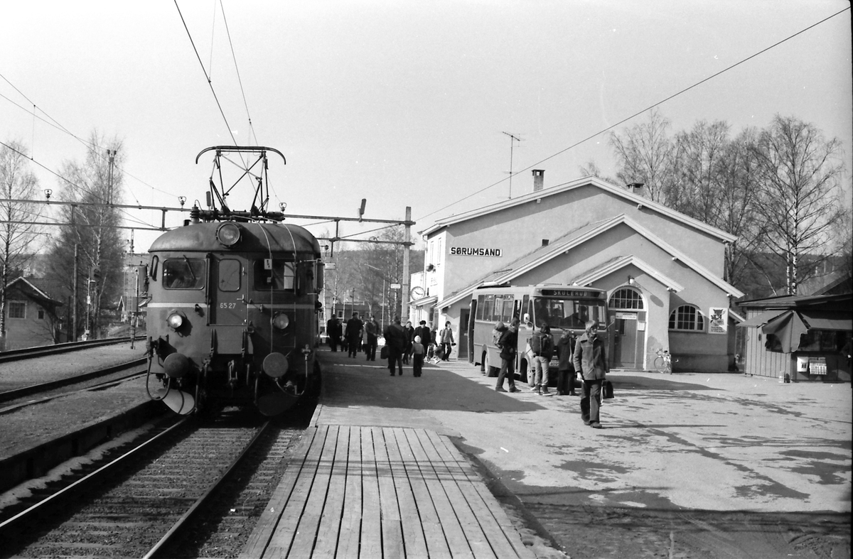 Persontog Oslo Ø - Årnes har stoppet på Sørumsand stasjon. Motorvogn BM 65B 27. Bytte til buss (Hølandsruta) for reisende til Skulerud. Bussen kjørte strekningen til den nedlagte Aurskog-Hølandbanen (Urskog-Hølandsbanen).