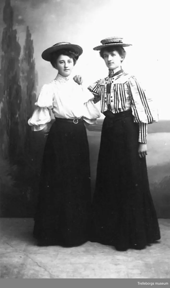 Två kvinnor i svart kjol, höghalsad blus och halmhatt.