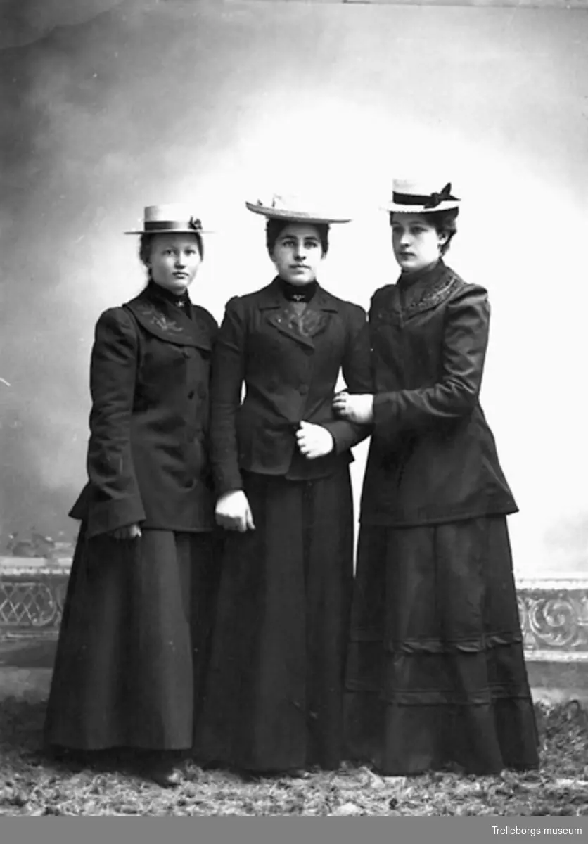 Tre kvinnor i svart tvådelad dräkt med broderad krage och halmhatt.