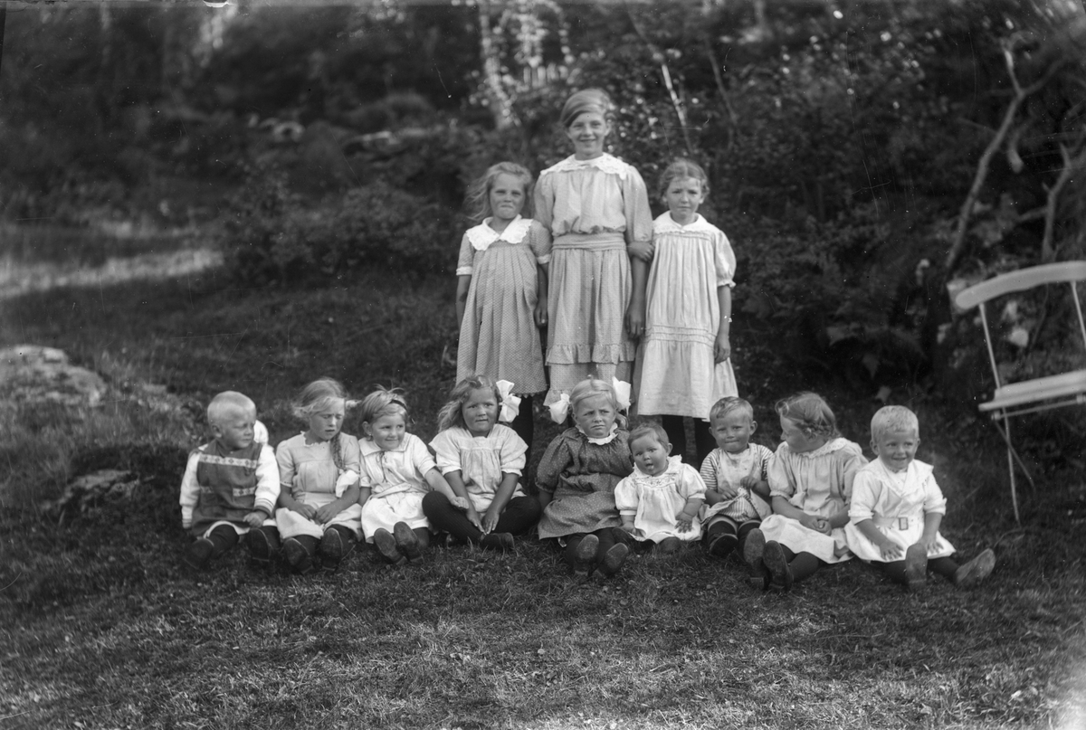 Gruppebilde av 12 barn, hvorav 10 jenter og 2 gutter. Bilde er tatt foran trær. En stol til høyre i bildet.