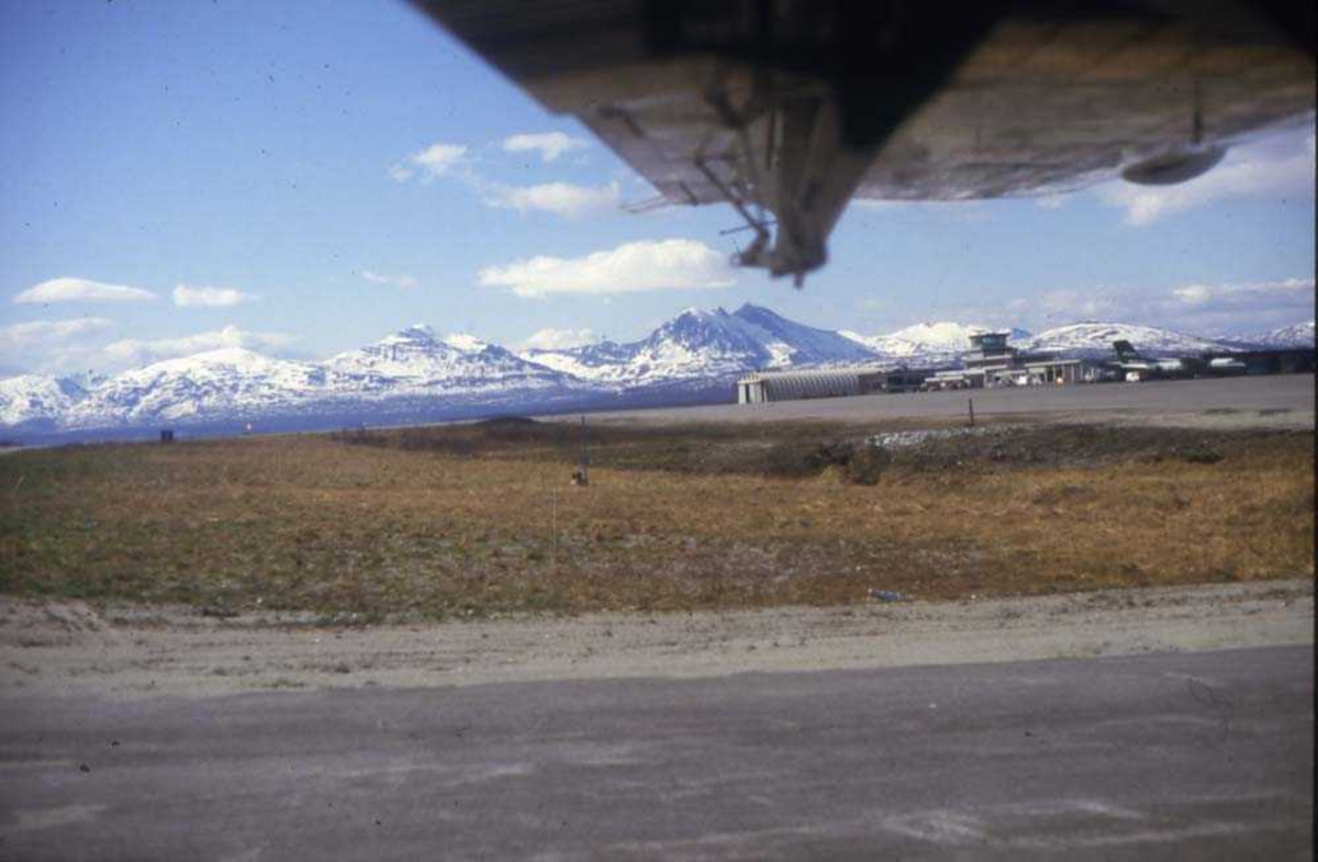 Lufthavn/flyplass. Et sted i nord. Et fly,  DHC-7-102 Dash7 fra Widerøe parkert. En underside av vingen med vinterkledde fjell i bakgrunnen.