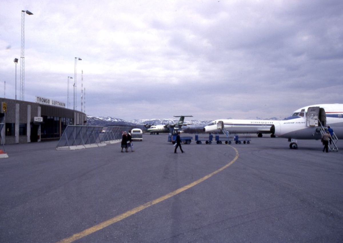Lufthavn (flyplass). Flere fly parkert på oppstillingsplassen (tarmac), to DC9 fra SAS og en DHC-7-102 fra Widerøe. Flere personer går ombord.