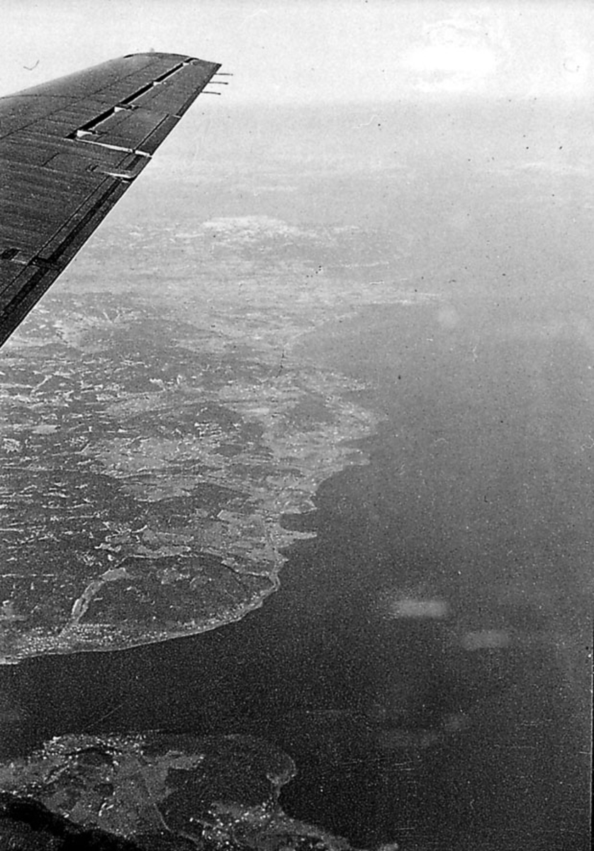 Luftfoto tatt fra fly, litt av vingen på flyet sees. Land med bebyggelse og hav under. 