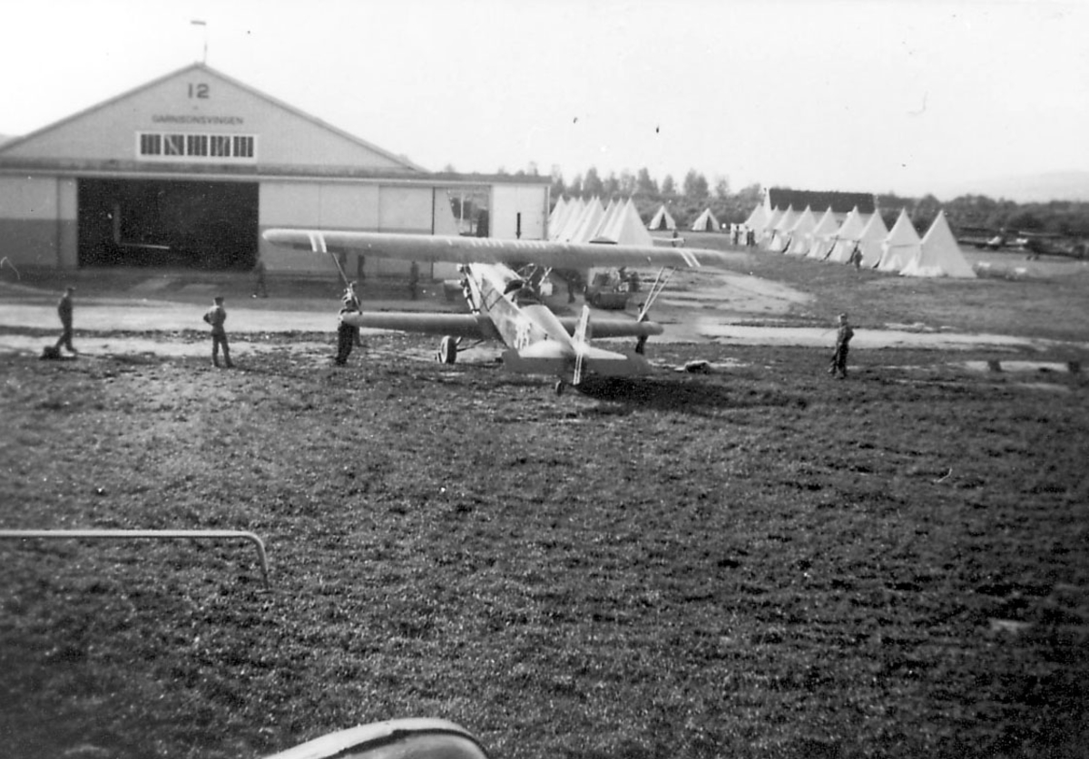 Åpen plass, lufthavn. Flere personer rundt ett fly på bakken, Fokker C.V.E., reg.nr. 315 Hangar bak med påskrift "Garnisonsvingen" og tallet 12. Flere telt er oppslått lenger bak.